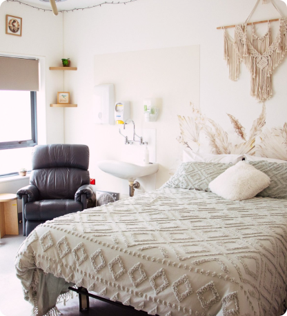 Cozy birthing suite at Mackay Birth Centre. There is with large bed with luxurious sheets and pillows, a comfortable padded chair, wall art, and a small sink. The room also has a window with a blind and a few decorative shelves with ornaments.